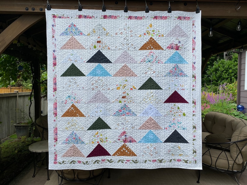 May include: A rectangular quilt featuring a repeating pattern of solid and floral-printed triangles arranged in a grid. The quilt has a white background with a floral border and is constructed with visible stitching throughout the fabric surface.