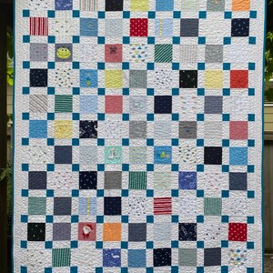 May include: A patchwork quilt featuring a grid of multi-colored fabric squares in various patterns, including stripes, polka dots, and cartoon graphics. The squares are separated by a teal blue lattice border and finished with a matching teal blue edge.
