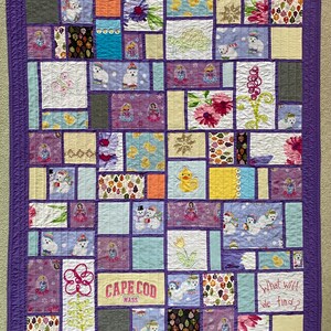 May include: A rectangular patchwork quilt featuring a purple border and various fabric squares. Patterns include polar bears, yellow ducks, floral prints, and a square with the embroidered text 'CAPE COD MASS.' and 'What will we find?'.