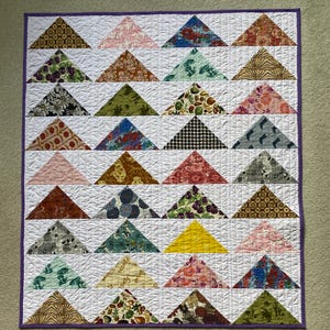 May include: A rectangular quilt featuring a grid of white fabric squares, each containing a single triangle made from various patterned fabrics in colors including yellow, green, red, blue, brown, and black, finished with a purple border.