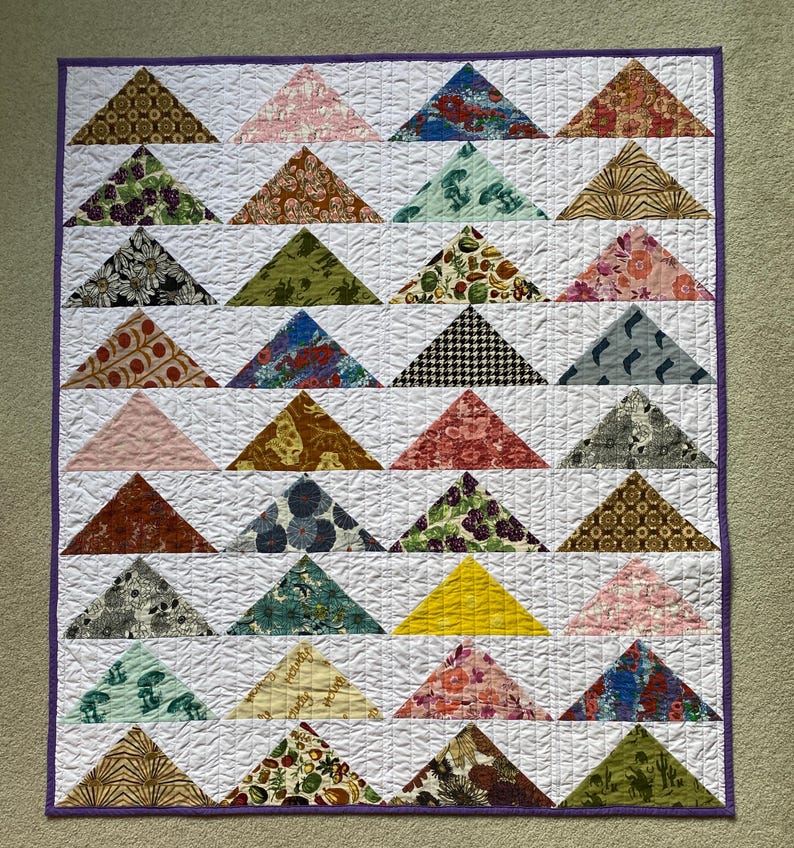 May include: A rectangular quilt featuring a grid of white fabric squares, each containing a single triangle made from various patterned fabrics in colors including yellow, green, red, blue, brown, and black, finished with a purple border.
