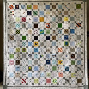 May include: A patchwork quilt featuring a grid of multi-colored fabric squares with various patterns, including animals, vehicles, and geometric shapes, separated by light gray sashing and bordered by a wide gray frame with a top hanging rod.