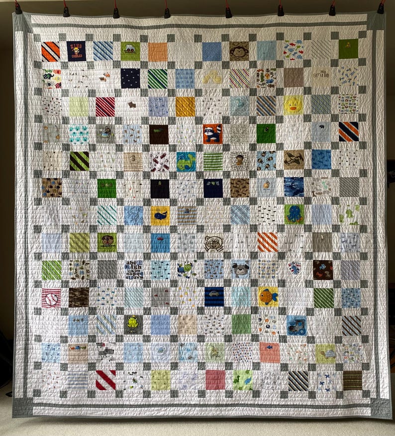 May include: A patchwork quilt featuring a grid of multi-colored fabric squares with various patterns, including animals, vehicles, and geometric shapes, separated by light gray sashing and bordered by a wide gray frame with a top hanging rod.