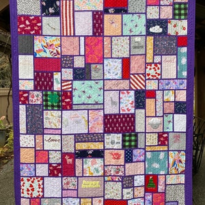 May include: A patchwork quilt featuring a variety of rectangular fabric blocks in diverse colors and patterns, including floral, animal, and geometric prints. The quilt is bordered in solid purple fabric and includes embroidered text like Grandma, loved, and happy human.