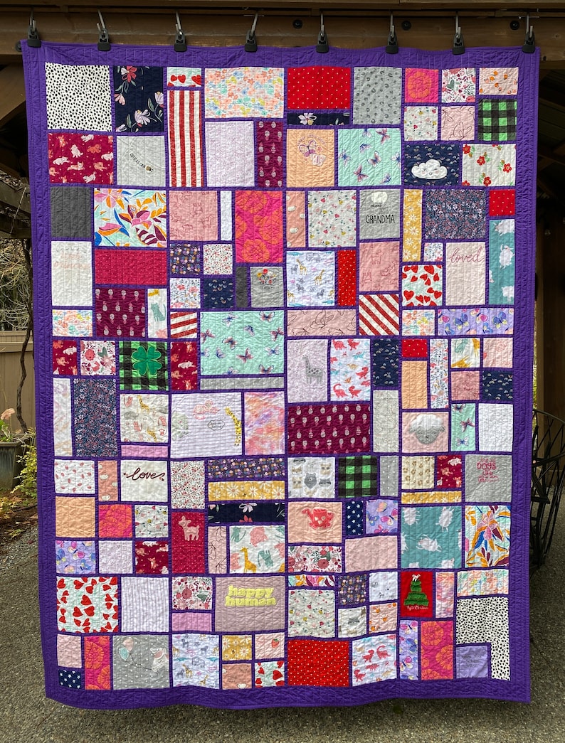 May include: A patchwork quilt featuring a variety of rectangular fabric blocks in diverse colors and patterns, including floral, animal, and geometric prints. The quilt is bordered in solid purple fabric and includes embroidered text like Grandma, loved, and happy human.