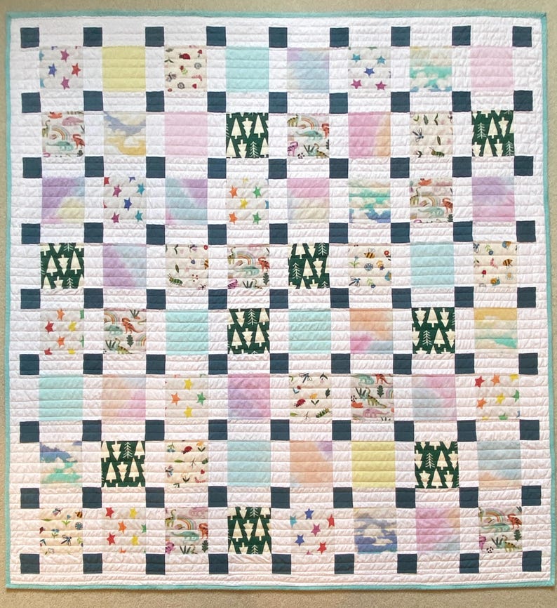 May include: A square patchwork quilt featuring a grid of alternating patterned and solid-colored fabric squares. The design includes dark blue sashing, white borders, and a light teal edge binding. Patterns include pine trees, stars, and dinosaurs.