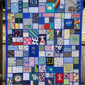 May include: A memory quilt featuring a patchwork of various fabric squares with diverse prints including a fire truck, NASA logo, a cow, rockets, and a Boichik Bagels graphic. The quilt is bordered by a solid blue fabric and stitched together.