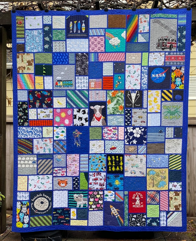May include: A memory quilt featuring a patchwork of various fabric squares with diverse prints including a fire truck, NASA logo, a cow, rockets, and a Boichik Bagels graphic. The quilt is bordered by a solid blue fabric and stitched together.