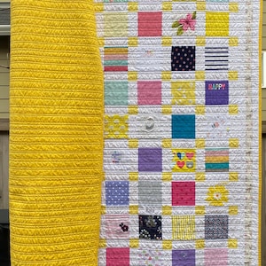 May include: A patchwork quilt featuring a grid of multi-colored fabric squares separated by white and yellow sashing. The quilt has a solid yellow backing with horizontal stitching. Some squares display patterns, floral prints, and the text HAPPY.