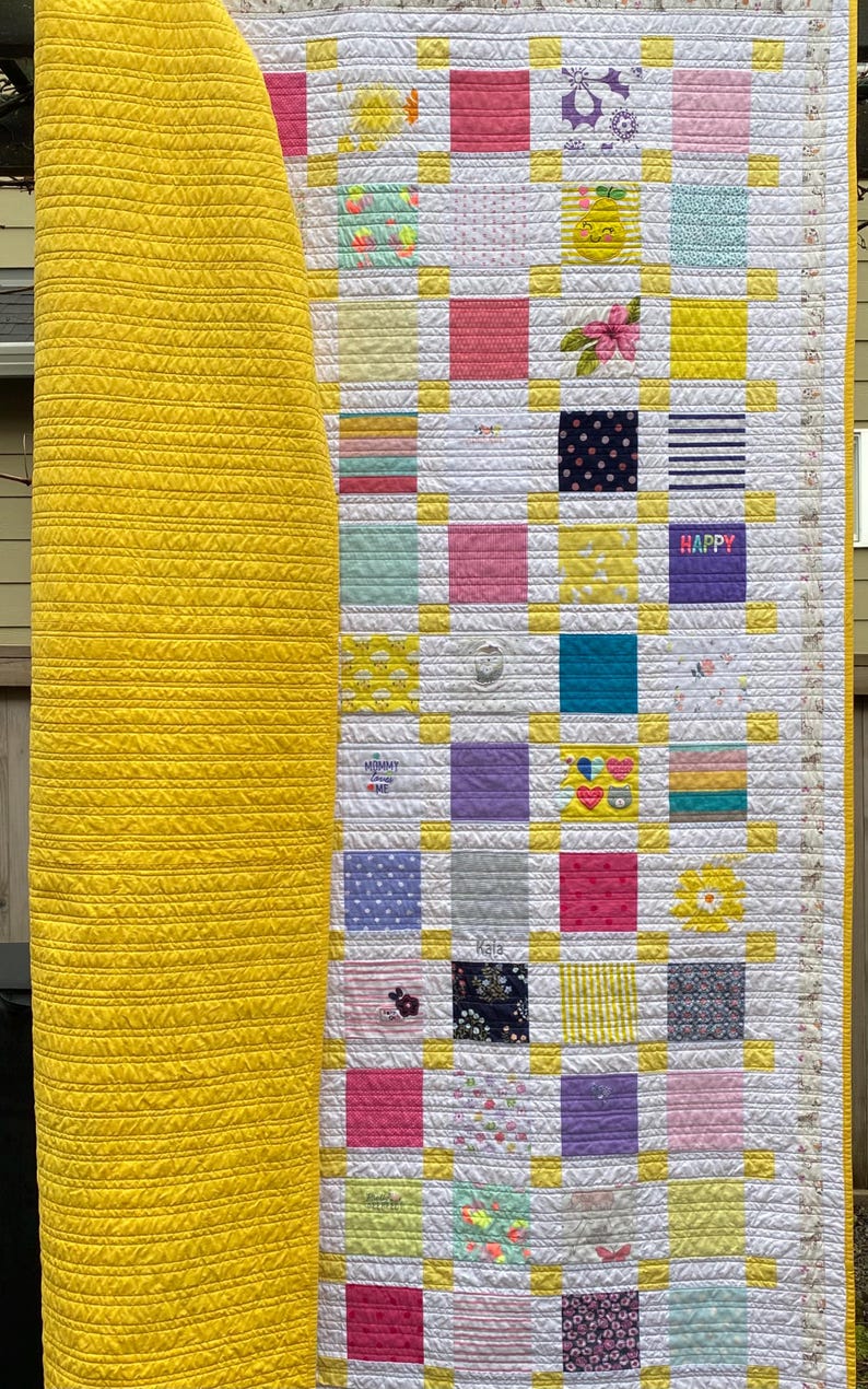 May include: A patchwork quilt featuring a grid of multi-colored fabric squares separated by white and yellow sashing. The quilt has a solid yellow backing with horizontal stitching. Some squares display patterns, floral prints, and the text HAPPY.