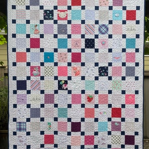 May include: A patchwork quilt featuring a grid of multi-colored fabric squares in shades of pink, blue, purple, and white, separated by small navy blue squares. The quilt includes various patterns and embroidered motifs like animals and flowers.