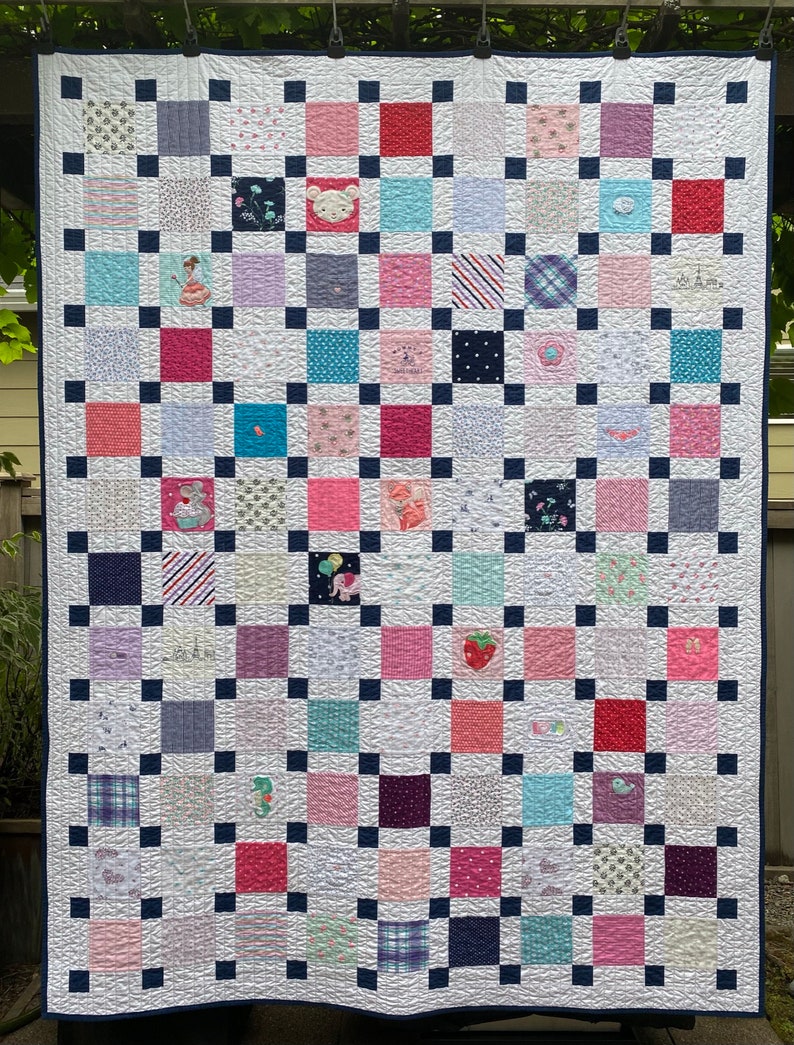 May include: A patchwork quilt featuring a grid of multi-colored fabric squares in shades of pink, blue, purple, and white, separated by small navy blue squares. The quilt includes various patterns and embroidered motifs like animals and flowers.