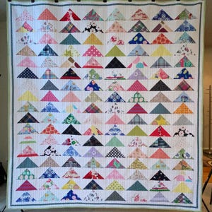 May include: A large rectangular quilt featuring a repeating pattern of multi-colored fabric triangles arranged in horizontal rows. The quilt has a white background with vertical quilting lines and a thin green border around the entire perimeter.