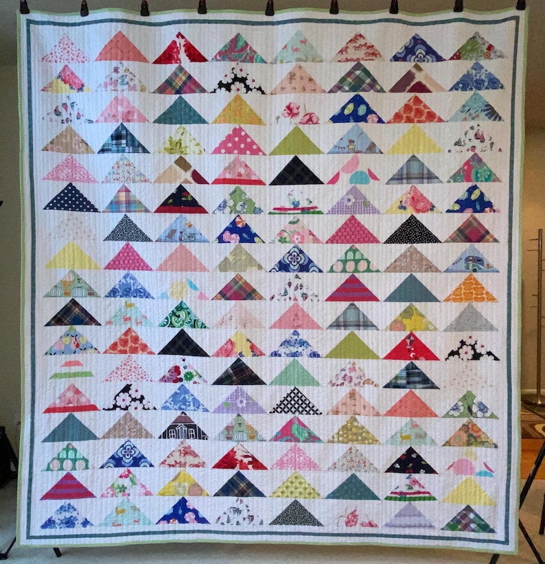 May include: A large rectangular quilt featuring a repeating pattern of multi-colored fabric triangles arranged in horizontal rows. The quilt has a white background with vertical quilting lines and a thin green border around the entire perimeter.