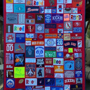 May include: A large rectangular memory quilt constructed from numerous colorful fabric patches featuring various sports team logos, athletic graphics, and text including names like Reed, team names, and motivational phrases, all stitched onto a red background.