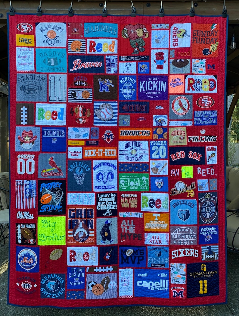 May include: A large rectangular memory quilt constructed from numerous colorful fabric patches featuring various sports team logos, athletic graphics, and text including names like Reed, team names, and motivational phrases, all stitched onto a red background.