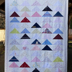 May include: A rectangular quilt featuring a grid of triangular fabric patches in various colors and patterns, including floral, plaid, and solid prints. The quilt has a white background with vertical and horizontal stitching and a thin border.