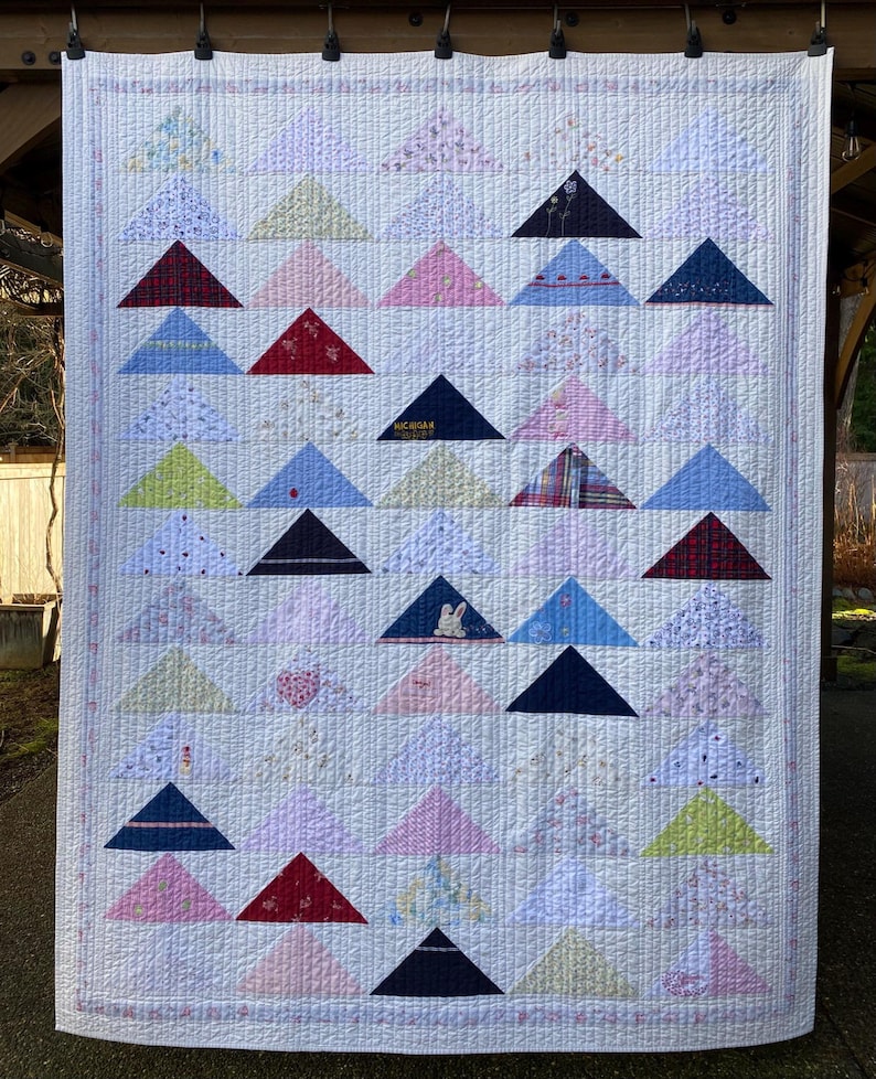 May include: A rectangular quilt featuring a grid of triangular fabric patches in various colors and patterns, including floral, plaid, and solid prints. The quilt has a white background with vertical and horizontal stitching and a thin border.