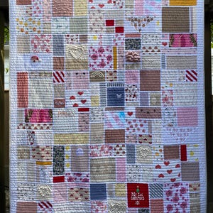May include: A patchwork quilt featuring a variety of rectangular fabric blocks in shades of pink, white, red, and grey. The quilt includes diverse patterns such as hearts, floral prints, stripes, and a red square with the text My 1st Christmas.
