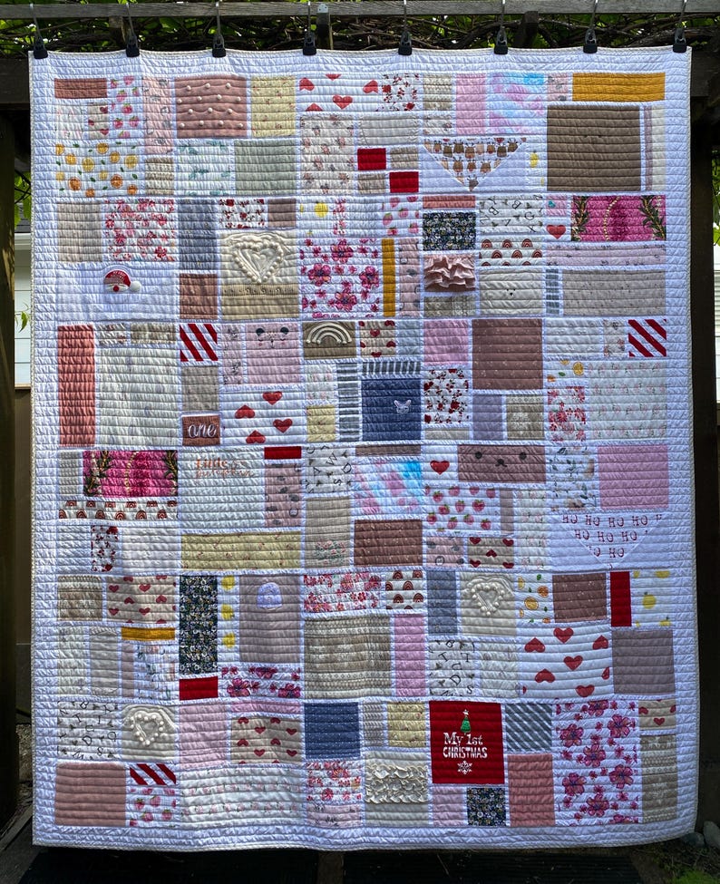 May include: A patchwork quilt featuring a variety of rectangular fabric blocks in shades of pink, white, red, and grey. The quilt includes diverse patterns such as hearts, floral prints, stripes, and a red square with the text My 1st Christmas.
