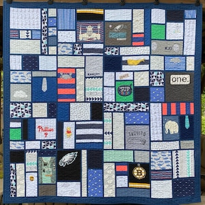 May include: A memory quilt constructed from various fabric squares and rectangles in shades of blue, gray, white, and green. The quilt features diverse graphic prints, including sports logos, animals, text like 'one.', 'Keegan', and 'Phillies', and geometric patterns, all stitched together with a dark blue border.