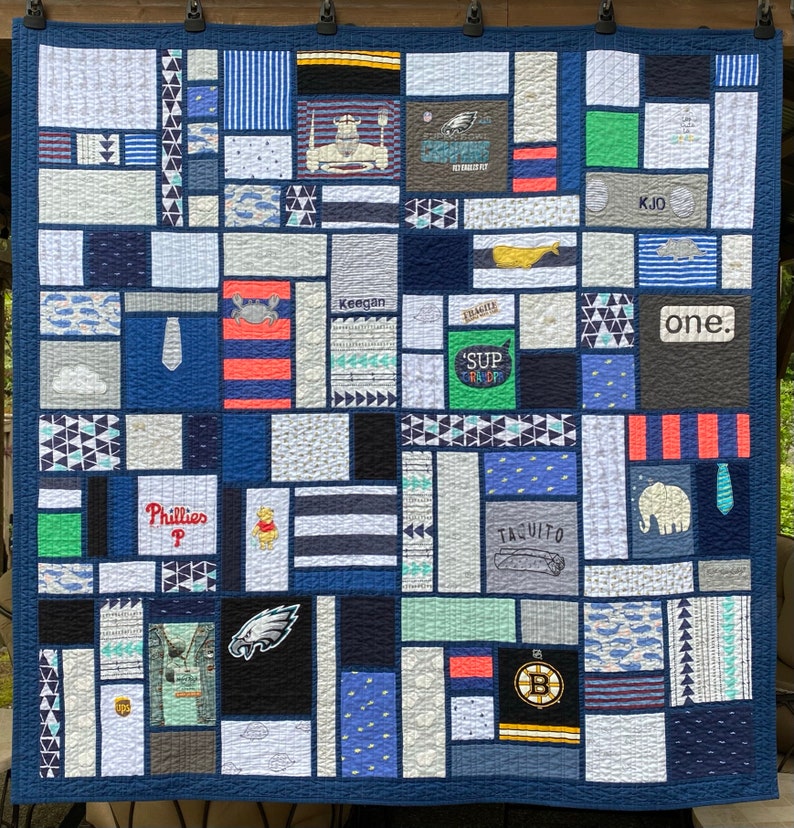 May include: A memory quilt constructed from various fabric squares and rectangles in shades of blue, gray, white, and green. The quilt features diverse graphic prints, including sports logos, animals, text like 'one.', 'Keegan', and 'Phillies', and geometric patterns, all stitched together with a dark blue border.
