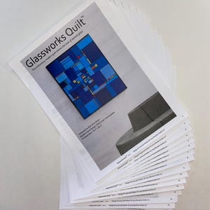 May include: A stack of white paper sheets with the title "Glassworks Quilt" and a colorful quilt design. The design mimics the look of stained glass. The finished size is 79.5 x 94.5 inches.