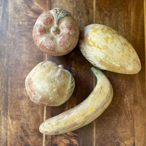 May include: Four decorative fruit sculptures are arranged on a wooden surface. The sculptures include a tomato, a mango, a banana, and a round fruit. The fruit sculptures are painted in shades of red, yellow, and brown. The fruit sculptures are arranged on a wooden surface.