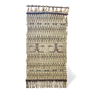 Puede incluir: Un tapiz de pared tejido con un patrón de diamantes en tonos beige, marrón y azul oscuro. La parte superior tiene una barra de madera con borlas y la parte inferior tiene flecos. El diseño incluye dos figuras estilizadas.