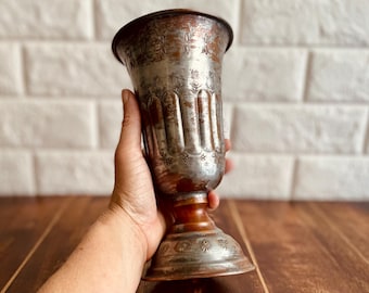 May include: An antique silver-colored goblet with a brown base and stem. The goblet has etched floral and star patterns. The cup is held in a hand against a white brick wall and a wooden surface.
