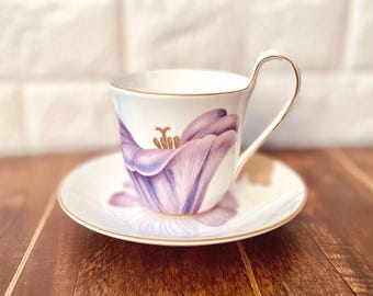 May include: White teacup and saucer set with a purple tulip design. The cup and saucer have gold trim. The tulip is painted in shades of purple and white. The handle of the cup is also gold.