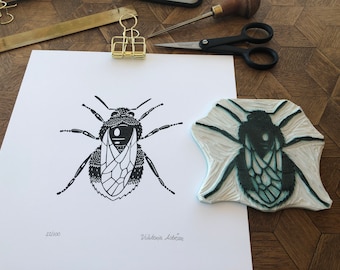 Puede incluir: Una impresión de linóleo en blanco y negro de una abeja con el número 22/100 y la firma del artista "Valetonia Lebon" en la esquina inferior derecha. La impresión está junto a un bloque de impresión de madera con un diseño de abeja.