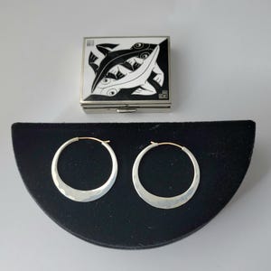 May include: A square metal box featuring a black and white graphic of two stylized fish on the lid, accompanied by a pair of circular silver hoop earrings with a hammered texture and thin gold-toned posts, displayed on a black semi-circular stand.