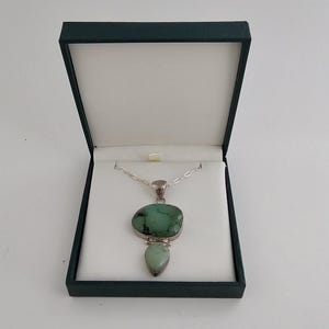 May include: A silver necklace featuring a pendant with two irregular, smooth green stones set in metal. The larger stone is rounded and dark green with black veining, while the smaller stone is a lighter, pale green teardrop shape. The necklace is displayed inside a dark blue, open jewelry box with a white interior lining.