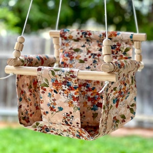 May include: A wooden baby swing with a floral patterned fabric seat. The swing is suspended from the ceiling by two white ropes.