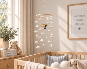 Puede incluir: Un móvil colgante para guardería con un marco circular de madera suspendido por cuerdas, adornado con múltiples nubes de fieltro blanco y un pequeño modelo de avión biplano amarillo. El móvil está diseñado para colgarse sobre una cuna.