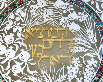 May include: Decorative artwork featuring a central circular design with gold Hebrew text. The circle is surrounded by white cut-out floral and fruit motifs, including pomegranates and wheat. The background has a repeating pattern of white leaves on a gray backdrop.