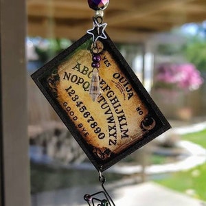 May include: A hanging Ouija board charm with a vintage aesthetic. The board features the alphabet, numbers, and the words "Yes" and "Good Bye." It is adorned with iridescent beads, a star charm, and a crystal pendant, creating a mystical and decorative piece.