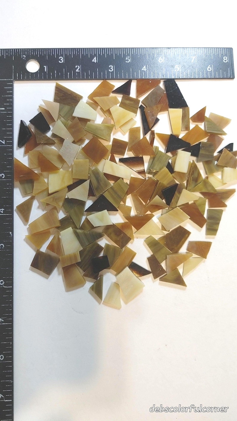 May include: A collection of irregularly shaped glass mosaic tiles in shades of brown, tan, and cream. A ruler is placed along the left side, with measurements in inches. The tiles are scattered on a white surface. The text "debscolorfulcorner" is in the bottom right corner.