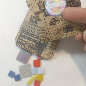 Puede incluir: Una peque&ntilde;a bolsa de papel con un dise&ntilde;o de peri&oacute;dico vintage, etiquetada como "Tiny bag of extras", se abre, derramando coloridas baldosas de mosaico. Las baldosas son de varias formas y colores, incluyendo rojo, azul, amarillo e iridiscente.