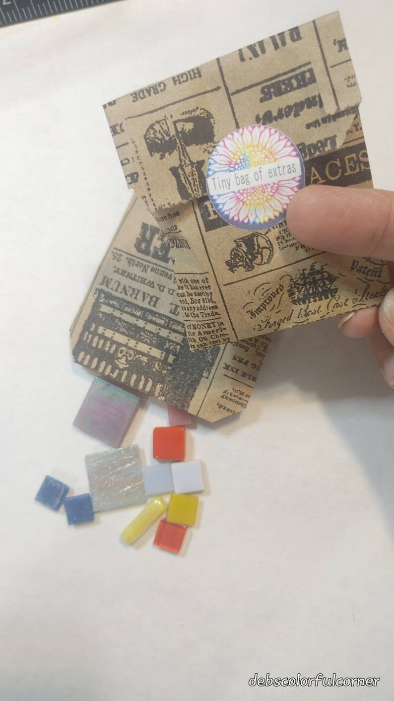 Puede incluir: Una peque&ntilde;a bolsa de papel con un dise&ntilde;o de peri&oacute;dico vintage, etiquetada como "Tiny bag of extras", se abre, derramando coloridas baldosas de mosaico. Las baldosas son de varias formas y colores, incluyendo rojo, azul, amarillo e iridiscente.