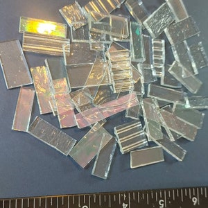 May include: A pile of iridescent glass tiles, some with a rainbow effect, on a blue surface with a black ruler measuring 6 inches.