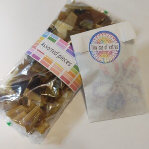 May include: Two clear plastic bags filled with assorted brown and tan glass pieces. One bag has a label that reads "Assorted pieces" in a colorful font. A small white paper bag with a floral design and the text "Tiny bag of extras" is also present.