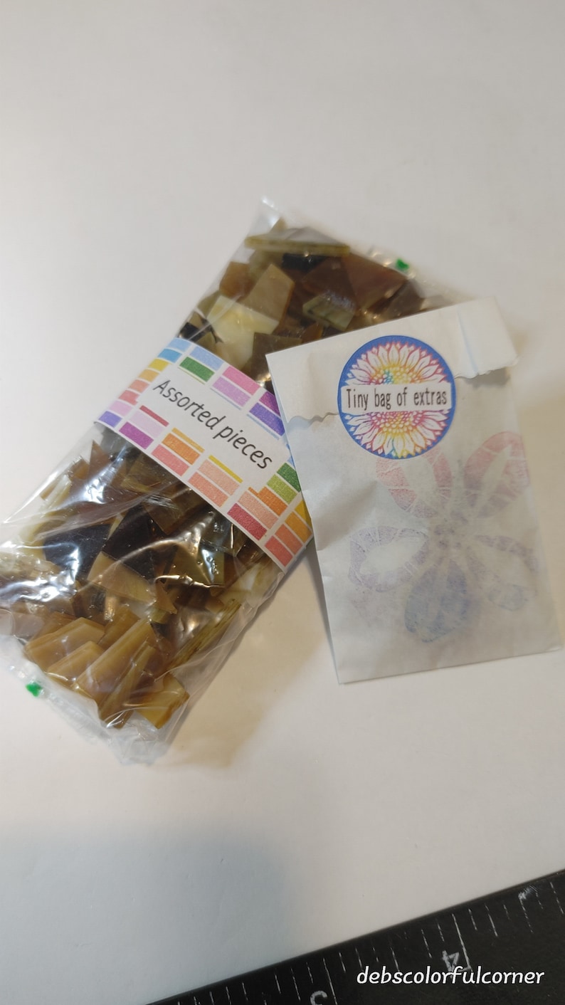 May include: Two clear plastic bags filled with assorted brown and tan glass pieces. One bag has a label that reads "Assorted pieces" in a colorful font. A small white paper bag with a floral design and the text "Tiny bag of extras" is also present.