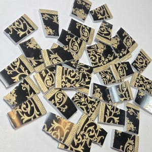 May include: A collection of irregular ceramic mosaic tiles featuring a black background with a cream-colored floral scroll pattern. The tiles are scattered above a black ruler showing measurements in inches, with the largest pieces around 1 inch wide.