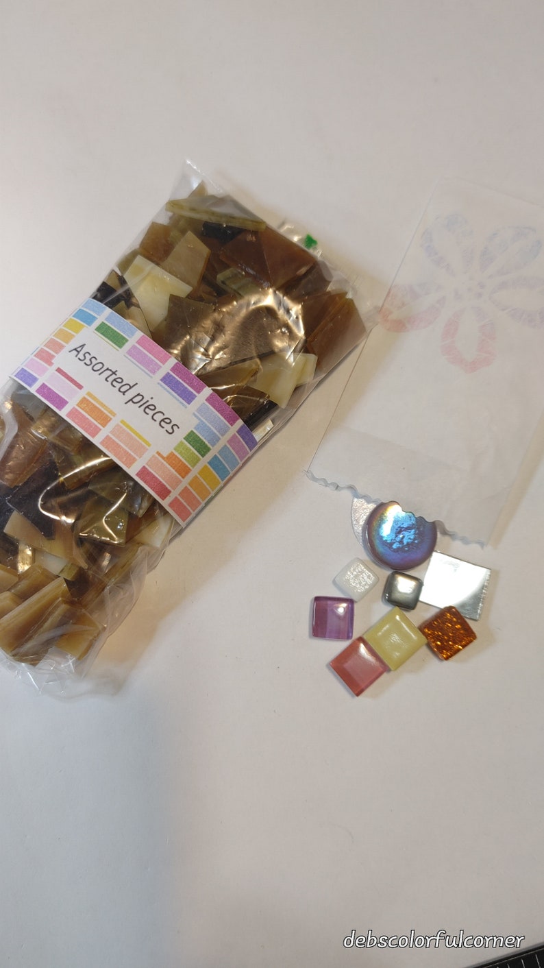May include: A clear plastic bag filled with assorted brown and tan mosaic pieces, next to a small bag with colorful square tiles. The label on the bag reads "Assorted pieces" in a rainbow-colored font.