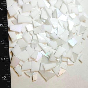 May include: A collection of small, irregular white mosaic tiles with iridescent surfaces. The tiles are scattered on a light background, with a black ruler along the left side. The tiles vary in shape and size.