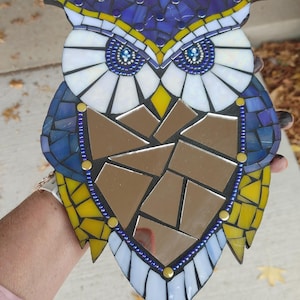 May include: A mosaic owl wall hanging with blue, yellow, and white glass tiles. The owl's eyes are made of iridescent blue beads. The owl's body is a mirror.