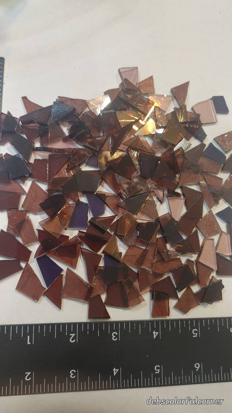 Puede incluir: Un conjunto de piezas de vidrio de colores de forma irregular, en tonos marrones, morados y dorados. Los fragmentos de vidrio est&aacute;n dispersos sobre una superficie blanca, con una regla negra para la escala. Piezas para arte de mosaico.
