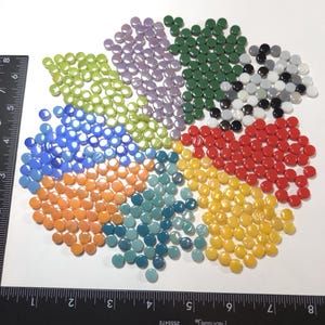 May include: A collection of small, round, multi-colored glass mosaic tiles arranged in a circular pattern on a white surface, accompanied by a black ruler showing measurements in inches to provide a clear sense of scale for the individual pieces.